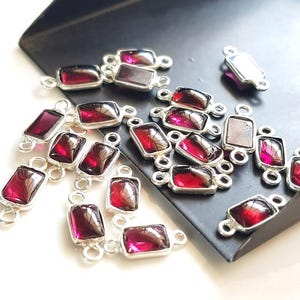 Puede incluir: Una colección de colgantes de piedras preciosas rojas con anillas de salto de plata. Los colgantes son de forma rectangular y presentan una piedra preciosa roja en el centro.