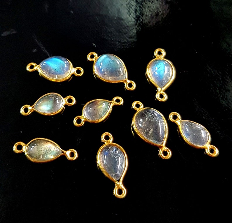 May include: Nine teardrop-shaped labradorite gemstone pendants with gold-colored bezels. The iridescent stones display blue and green hues. Each pendant has small loops on either end for attaching to jewelry. The pendants are displayed on a black surface.