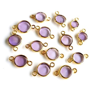 Puede incluir: Una colección de conectores de bisel dorados con inserciones redondas de vidrio morado claro. Los conectores tienen pequeños bucles a cada lado para sujetar. Las piezas están dispersas sobre una superficie blanca.