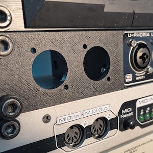 Pode incluir: Close-up de um painel preto com duas grandes aberturas circulares, parte de um sistema de interface de áudio montado em rack. O painel é fixado com parafusos e fica acima de outros equipamentos de áudio, incluindo uma interface U-PHORIA UMC404HD e uma unidade ME 30R.