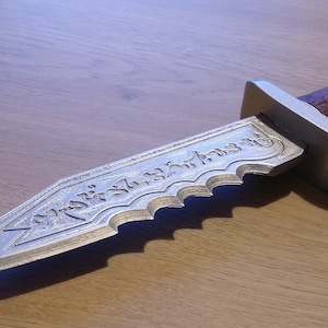 Peut inclure: Un poignard argenté avec une lame ornée et un manche marron texturé. La lame présente du texte gravé et un bord dentelé. Le manche a l'aspect du bois. Le poignard est posé sur une surface en bois.