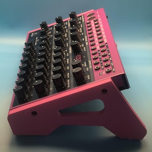 May include: A bright pink synthesizer featuring many black knobs and buttons. The electronic music instrument has a series of small buttons and input jacks on the right. The synthesizer is angled upwards on a pink stand.