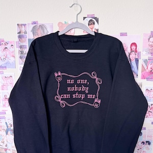 May include: Black crewneck sweatshirt with the text "no one, nobody can stop me" in pink script. The text is framed by a decorative pink border with bunny illustrations. The sweatshirt is hanging on a gray hanger.