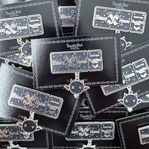 Può includere: Un set di spille in smalto nero e argento con un design a biglietto con il testo "Eroi ordinari" e "Ammetti uno". Le spille sono esposte su cartoncino nero con un motivo a catena.