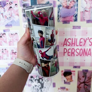 May include: A stainless steel tumbler featuring a comic book style design with images of Spider-Man. The tumbler has a clear lid and is decorated with various panels of the superhero in action. The background includes posters and the text "ASHLEY'S PERSONA".