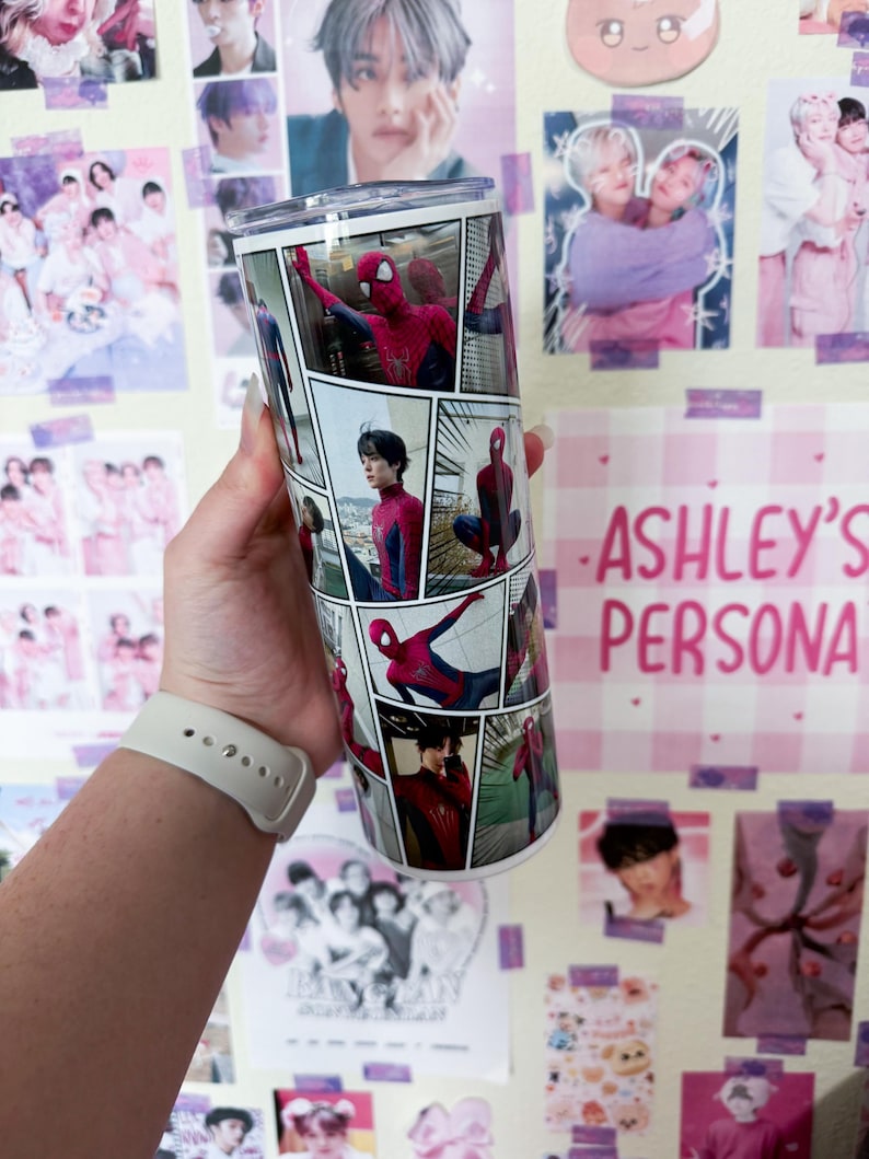 May include: A stainless steel tumbler featuring a comic book style design with images of Spider-Man. The tumbler has a clear lid and is decorated with various panels of the superhero in action. The background includes posters and the text "ASHLEY'S PERSONA".
