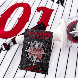 May include: A black and white baseball jersey with the number "01" in red on the front. The jersey has the word "JEONGIN" in black above the number. A black and white striped lanyard with a red star is hanging from the jersey. A white and red light-up stick is on the right side of the jersey. A black and silver pin with the words "Stray Kids" and "Dominate" in red is on the left side of the jersey.