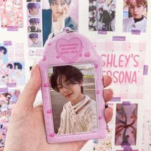May include: A pink Kpop Idol photo holder with a photo of a young man. The holder has a heart-shaped design with the words "EMOTIONAL SUPPORT KPOP IDOL". The background features Kpop posters.