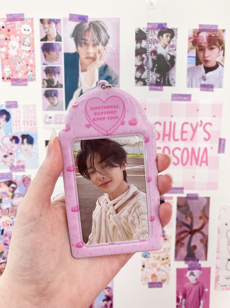 May include: A pink Kpop Idol photo holder with a photo of a young man. The holder has a heart-shaped design with the words "EMOTIONAL SUPPORT KPOP IDOL". The background features Kpop posters.