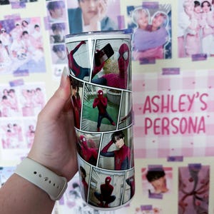 May include: A stainless steel tumbler featuring a comic book style design with images of Spider-Man. The tumbler has a silver lid and the text "ASHLEY'S PERSONA" in pink. The background includes various posters.