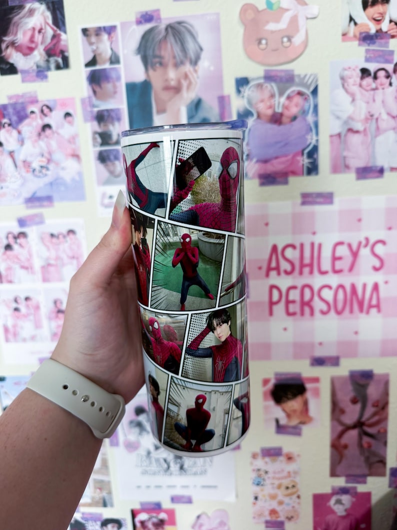 May include: A stainless steel tumbler featuring a comic book style design with images of Spider-Man. The tumbler has a silver lid and the text "ASHLEY'S PERSONA" in pink. The background includes various posters.