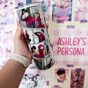 May include: A white tumbler featuring a comic strip design with images of Spider-Man. The tumbler has a clear lid and is held against a wall decorated with posters and the text "ASHLEY'S PERSONA".