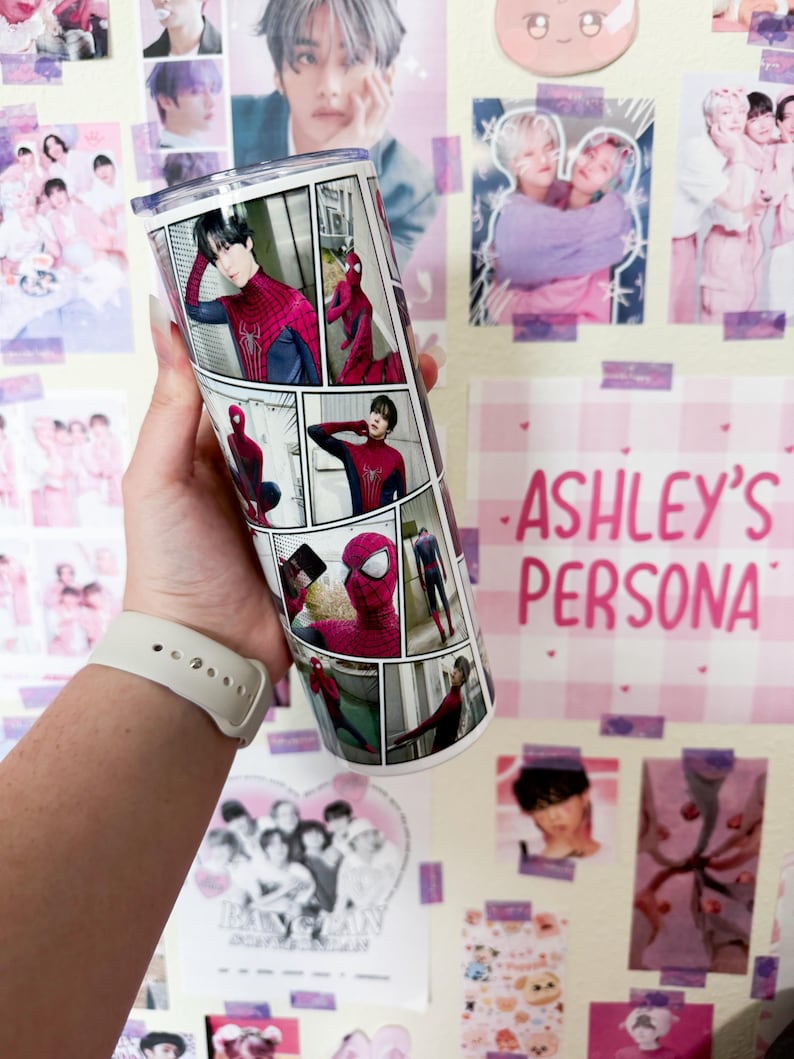 May include: A white tumbler featuring a comic strip design with images of Spider-Man. The tumbler has a clear lid and is held against a wall decorated with posters and the text "ASHLEY'S PERSONA".