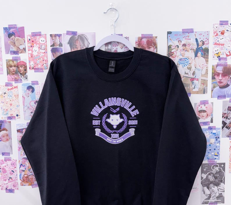 May include: Black crewneck sweatshirt with a purple graphic that reads "Villainsville" with a crest design. The sweatshirt is hanging on a white hanger against a wall decorated with various posters.
