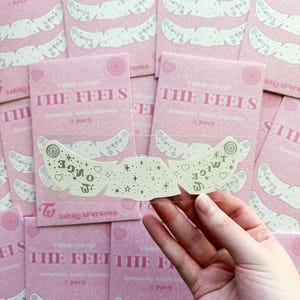 May include: Pink temporary tattoo packs with the text "THE FEELS" and "2 pack". The tattoos are white and feature a design with stars, hearts, and the word "TWICE".