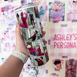 May include: A stainless steel tumbler featuring a comic strip design with images of Spider-Man and a person in a red and black suit. The tumbler is cylindrical and has a silver lid. The background includes posters and the text "ASHLEY'S PERSONA".