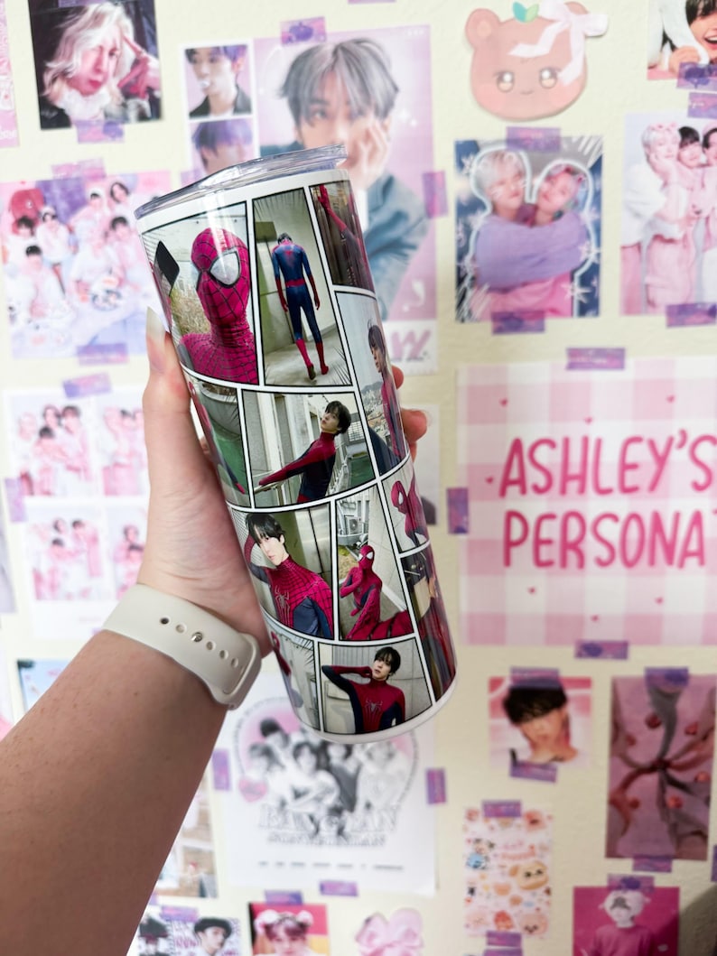 May include: A stainless steel tumbler featuring a comic strip design with images of Spider-Man and a person in a red and black suit. The tumbler is cylindrical and has a silver lid. The background includes posters and the text "ASHLEY'S PERSONA".