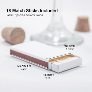 a close-up view of a matchbox with 18 matchsticks, including white-tipped and natural wood-ended matches. The matchbox is placed on a white surface, with a blurred background that includes a wine glass and a cork. The image provides measurements for the matchbox, including its length, height, and width.