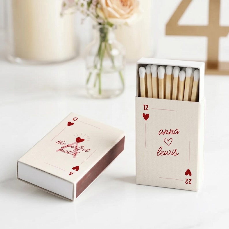 May include: Two matchboxes with a cream-coloured background. One box is closed and has the text "the perfect match" with a heart design. The other box is open, revealing wooden matches with white tips. The open box has the names "anna lewis" and a heart.