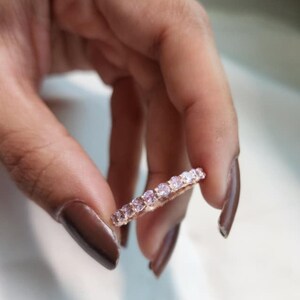 May include: A rose gold eternity band featuring a continuous row of round, light pink gemstones set in a shared prong setting. The ring displays a metallic rose gold finish and a series of uniform, faceted pink stones that span the entire circumference.