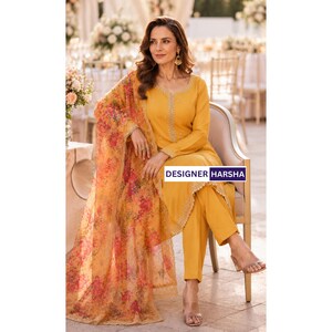 Puede incluir: Un conjunto de t&uacute;nica y pantalones color amarillo mostaza con bordados intrincados a lo largo del escote y el dobladillo. El atuendo incluye un dupatta transparente con estampado floral en tonos naranja, rosa y amarillo. El texto DESIGNER HARSHA es visible.