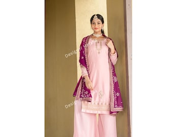 Puede incluir: Un conjunto kurta rosa claro que incluye una túnica de manga larga con bordados dorados en el cuello y el dobladillo, junto con pantalones anchos. Un dupatta morado oscuro con bordados florales dorados cubre los hombros. Texto Designer Harsha.