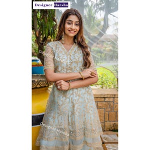 May include: A woman wearing a light blue embroidered dress with gold accents. The dress has a V-neckline and a flared skirt. The woman is standing in front of a green background.