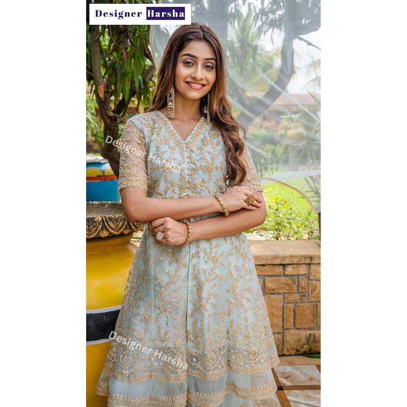 May include: A woman wearing a light blue embroidered dress with gold accents. The dress has a V-neckline and a flared skirt. The woman is standing in front of a green background.