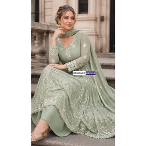 May include: A sage green long-sleeved dress featuring a V-neckline, intricate silver embroidery, and reflective mirror embellishments throughout the bodice and skirt. The garment includes a matching sheer dupatta and the text DESIGNER HARSHA.