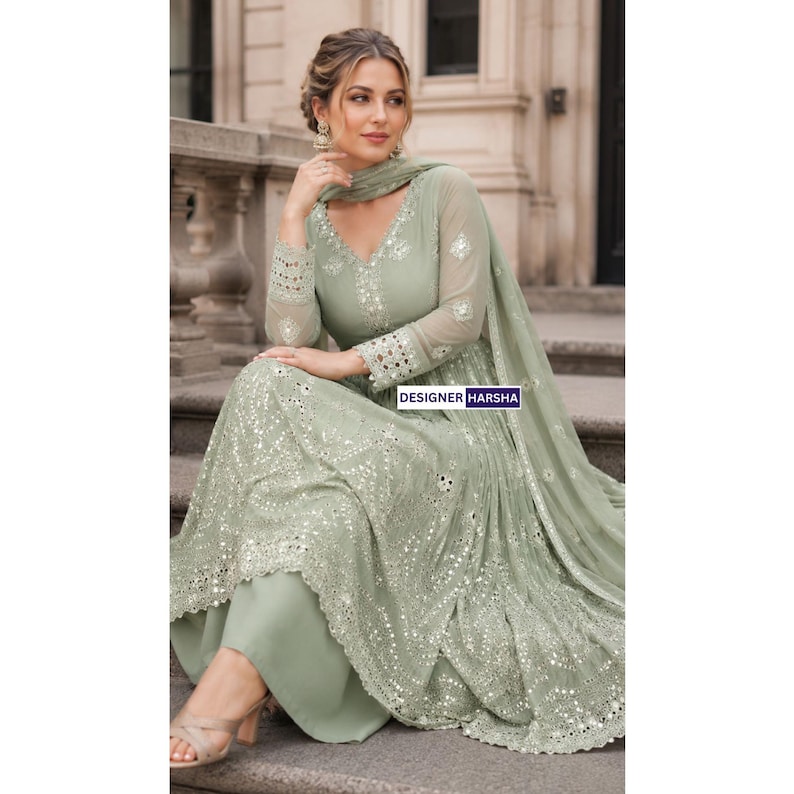 May include: A sage green long-sleeved dress featuring a V-neckline, intricate silver embroidery, and reflective mirror embellishments throughout the bodice and skirt. The garment includes a matching sheer dupatta and the text DESIGNER HARSHA.
