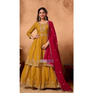 May include: Mustard yellow long-sleeved dress with intricate gold embroidery and embellishments. A vibrant red dupatta is draped over the shoulder. The dress features a tiered skirt and is labeled "DESIGNER HARSHA".