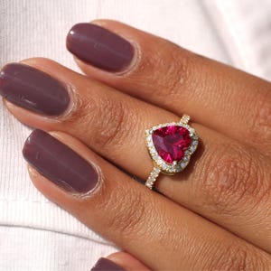 Puede incluir: Un anillo de oro con una piedra preciosa roja oscura en forma de corazón, rodeada por un halo de piedras claras más pequeñas. El anillo está en un dedo con esmalte de uñas morado oscuro. La banda del anillo también está adornada con pequeñas piedras claras.