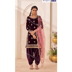 May include: A deep purple velvet salwar kameez set with gold embroidery. The outfit includes a long-sleeved tunic, matching pants, and a sheer pink dupatta. The text "Designer Harsha" is visible.