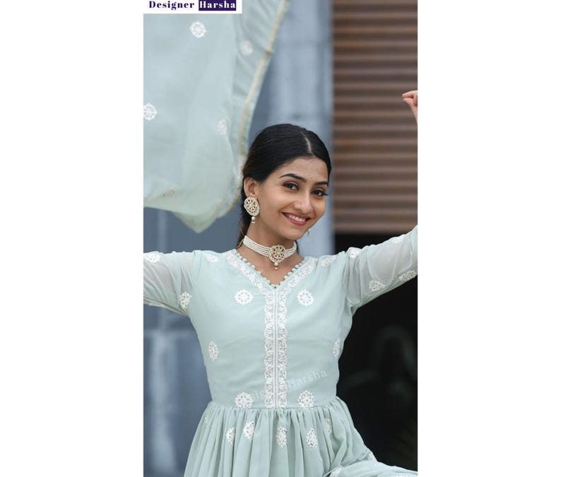 May include: A woman wearing a light blue embroidered dress with white embroidery. She is wearing a pearl necklace and earrings. The dress has a ruffled hem and a V-neckline. The woman is smiling and looking at the camera.
