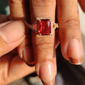 Könnte beinhalten: Ein goldener Ring mit einem großen, rechteckigen, roten Edelstein. Der Edelstein ist von einem Kranz aus kleineren, klaren Steinen umgeben. Der Ring hat ein einzigartiges Design mit dreieckigen Akzenten auf beiden Seiten des Hauptsteins.