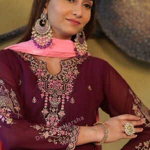 May include: A woman wearing a maroon and pink embroidered dress with gold jewelry. The dress has a deep V-neckline and a long, flowing skirt. The woman is wearing a pink dupatta over her head and shoulders. The dress is decorated with intricate embroidery and sequins.