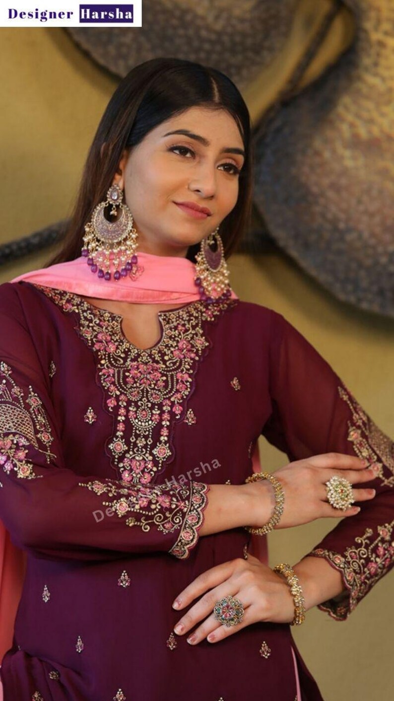 May include: A woman wearing a maroon and pink embroidered dress with gold jewelry. The dress has a deep V-neckline and a long, flowing skirt. The woman is wearing a pink dupatta over her head and shoulders. The dress is decorated with intricate embroidery and sequins.