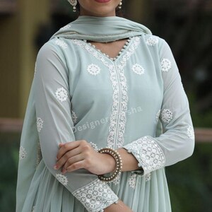 May include: A light blue dress with white embroidery. The dress has a V-neckline and long sleeves. The dress is made of a sheer fabric and has a flowing silhouette. The dress is decorated with white beads and sequins. The dress is worn by a woman with dark hair and a light complexion. The woman is standing in front of a green background.