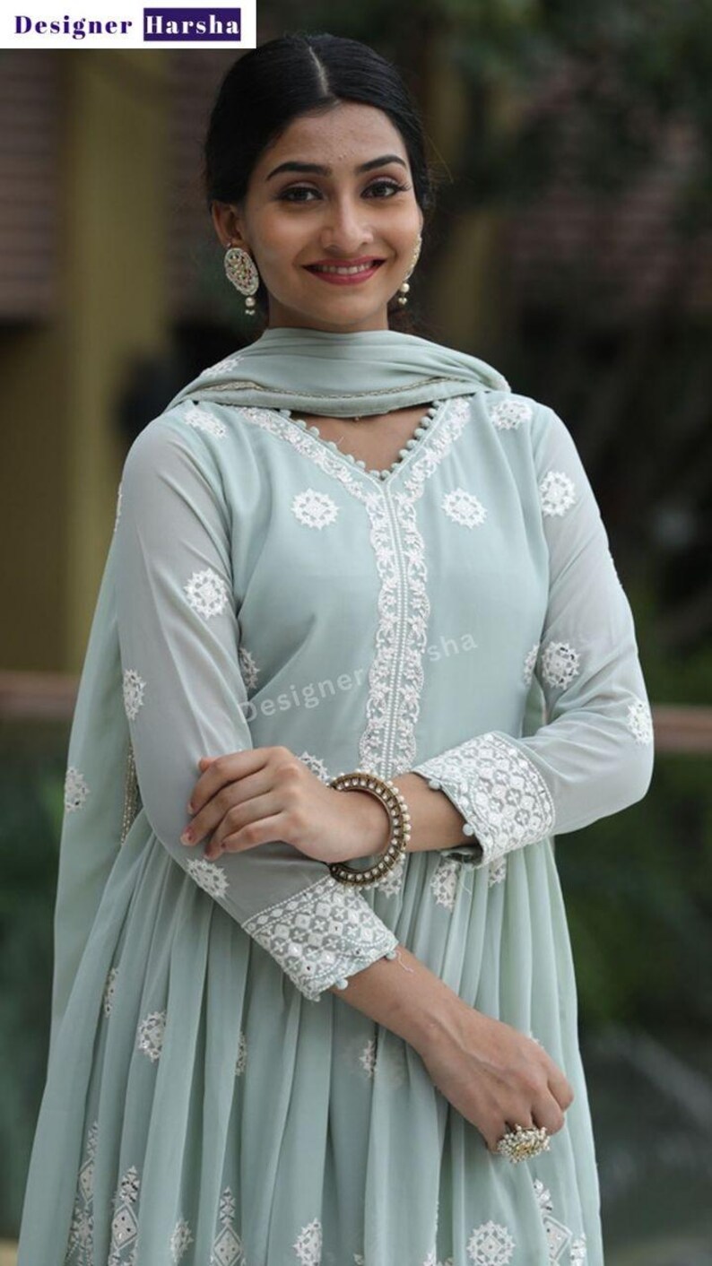 May include: A light blue dress with white embroidery. The dress has a V-neckline and long sleeves. The dress is made of a sheer fabric and has a flowing silhouette. The dress is decorated with white beads and sequins. The dress is worn by a woman with dark hair and a light complexion. The woman is standing in front of a green background.