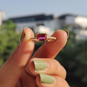 May include: A gold ring with a large, rectangular, deep pink gemstone. The band is decorated with small, clear accent stones. The ring is held up against a blurred background.