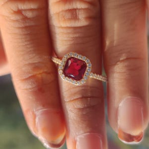 May include: A gold ring featuring a square, deep red gemstone surrounded by a halo of smaller, clear stones. The ring's band is also adorned with small, clear stones. The ring is on a finger.