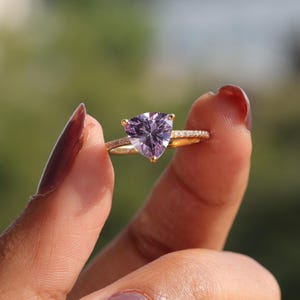 May include: A gold ring featuring a triangular, light purple gemstone. The ring's band is also adorned with small, clear stones. The ring is held up against a blurred green background, showcasing the ring's design and color.