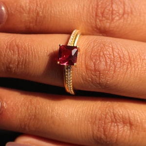 Könnte beinhalten: Ein goldener Ring mit einem quadratischen, tiefroten Edelstein. Das Band ist geteilt, eine Seite mit kleinen, klaren Steinen besetzt. Der Ring wird vor einem warmen, neutralen Hintergrund präsentiert, der die lebendige Farbe des Edelsteins hervorhebt.