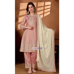 May include: A blush pink salwar kameez ensemble with intricate embroidery. The outfit includes a long-sleeved tunic, matching pants, and a cream-colored shawl. The text "DESIGNER HARSHA" is visible.