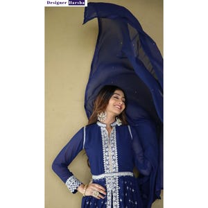 May include: A woman wearing a navy blue dress with white embroidery and a matching blue dupatta. The dress has a V-neckline and long sleeves. The dupatta is draped over her head and shoulders.