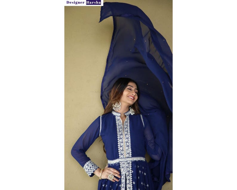May include: A woman wearing a navy blue dress with white embroidery and a matching blue dupatta. The dress has a V-neckline and long sleeves. The dupatta is draped over her head and shoulders.