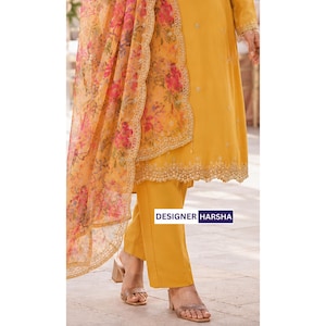 Puede incluir: Una t&uacute;nica de color amarillo mostaza con dobladillo festoneado y pantalones a juego, combinada con una dupatta naranja transparente con un estampado floral en rosa y naranja. La prenda incluye el texto DESIGNER HARSHA en un recuadro azul.