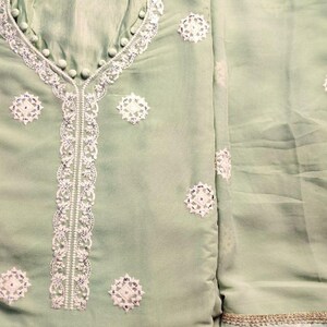 May include: A light sage green tunic featuring white embroidered floral motifs and a central vertical lace panel. The neckline is accented with small fabric-covered buttons. The text DESIGNER HARSHA is visible at the top. The fabric has a subtle sheen.