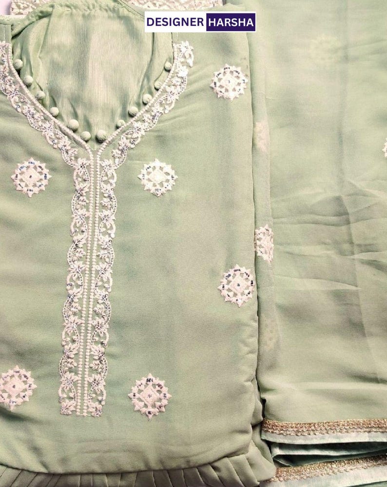May include: A light sage green tunic featuring white embroidered floral motifs and a central vertical lace panel. The neckline is accented with small fabric-covered buttons. The text DESIGNER HARSHA is visible at the top. The fabric has a subtle sheen.