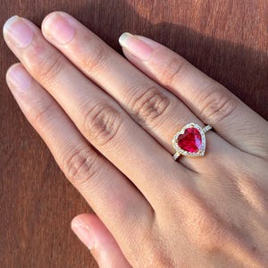 以下が含まれることがあります： ハート型の赤い宝石と小さな透明な石で囲まれた金の指輪。指輪は、木製の背景に対して、人の指にあります。指輪のバンドも金です。